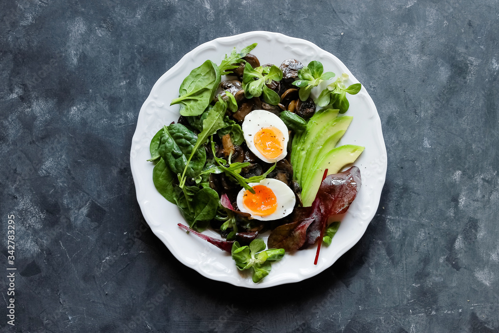 Healthy breakfast eggs, avocado, mushrooms and fresh greens arugula, spinach on a dark plate top view