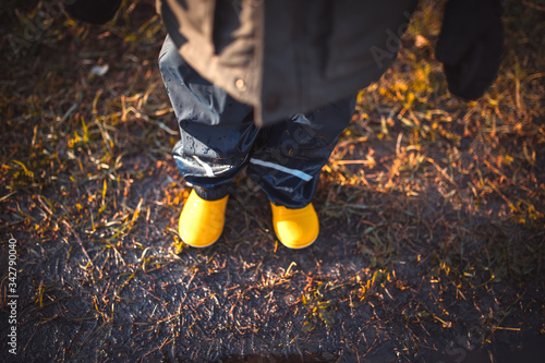 a child in yellow rubber boots, in a jacket, stands on wet ground. Spring. View from above.sunny weather