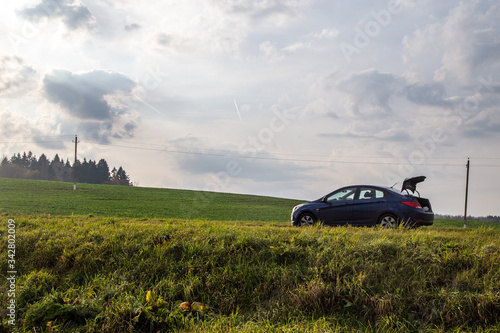 Car standing in the fields. Sky and clouds in background. Forest, trees on a hill. Green grass, yellow flowers. 
