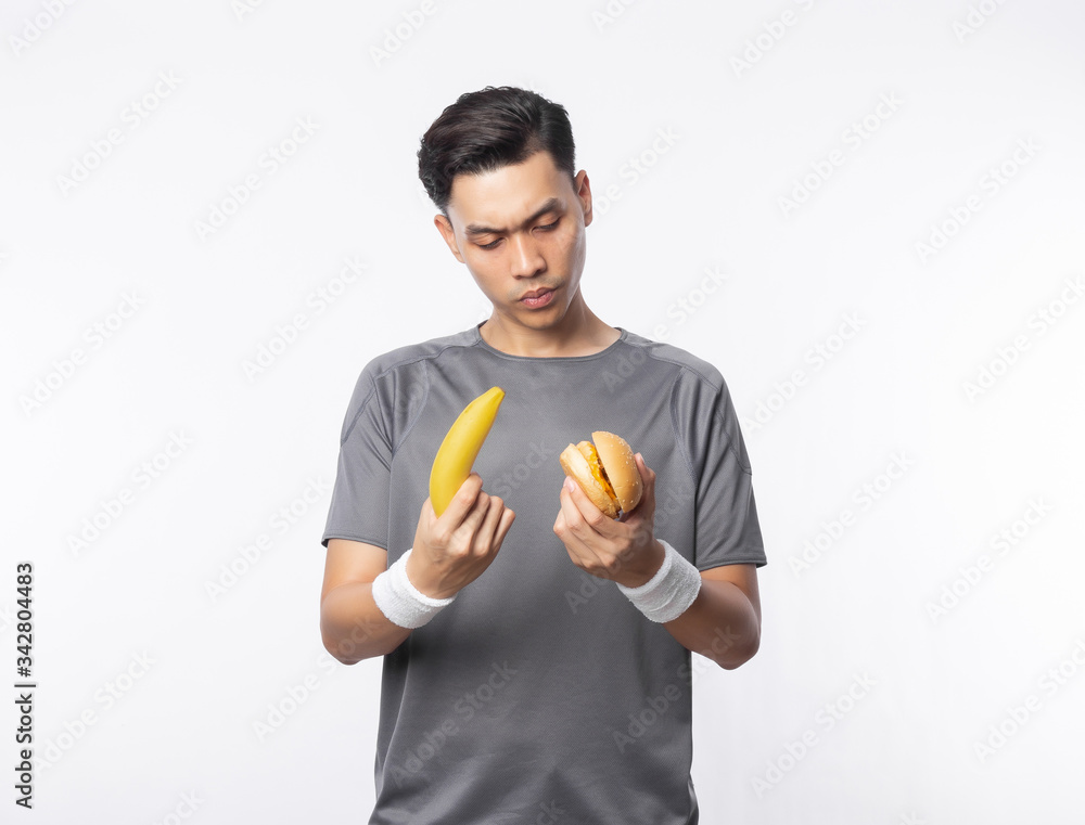 Young asian man in sport outfits holding banana and hamburger with doubts face isolated on white background. Young asian man in sport outfits holding banana and hamburger with doubts face isolated on white background.