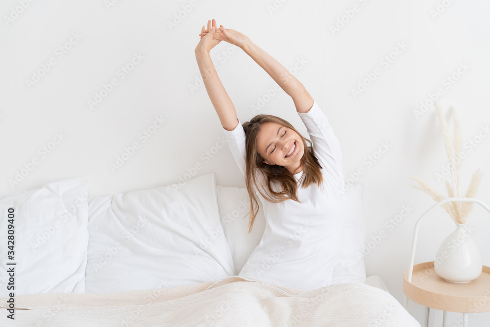 Young positive girl sitting on bed after waking up in morning, stretching arms up and left to get more energy and strength, smiling happily Young positive girl sitting on bed after waking up in morning, stretching arms up and left to get more energy and strength, smiling happily