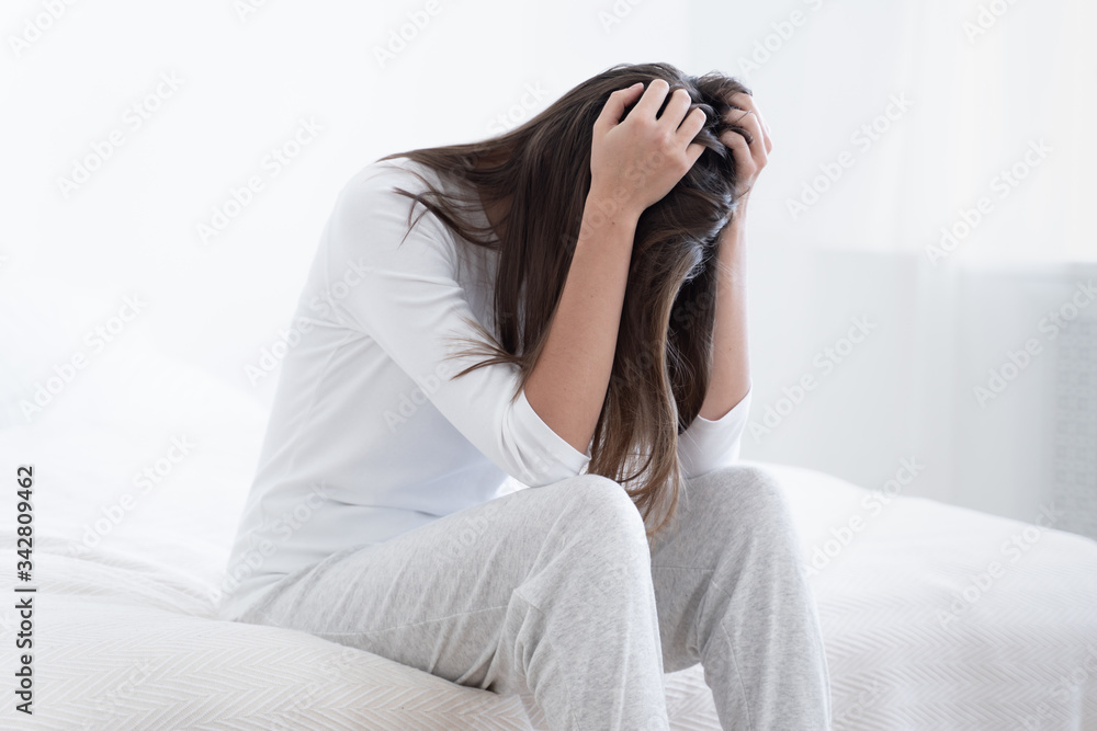 Woman spending afternoon in bedroom, suffering from insomnia and severe headache, holding her head, sitting on bed, looking desperate and exhausted Woman spending afternoon in bedroom, suffering from insomnia and severe headache, holding her head, sitting on bed, looking desperate and exhausted
