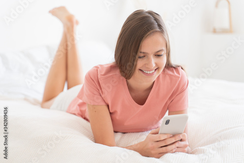 Young lady smiling happily while chatting with friends or boyfriend in text messages, lying on white bed, dressed in home clothes