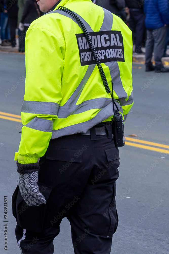 A male medical first responder walks along a street alone with people standing in the distance. The officer is wearing a bright yellow coat with grey reflective bands, black pants and a radio. A male medical first responder walks along a street alone with people standing in the distance. The officer is wearing a bright yellow coat with grey reflective bands, black pants and a radio.