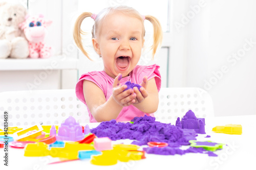 Girl plays kinetic sand in quarantine. Blond beautiful girl smiles and plays with purple sand on a white table.