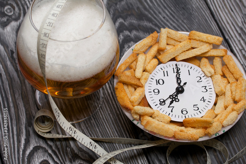 A clock showing seven o'clock in the evening. Lie in a cup with crackers. Near a glass of beer and a measuring tape. Against the background of brushed pine boards in black. World No Diet Day.