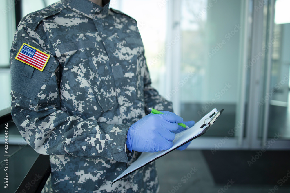 Fototapeta premium American soldier in camouflage uniform with rubber gloves, mask guarding in front of hospital's door and controlling who gets in and out. USA military helping in fight against coronavirus or COVID-19.