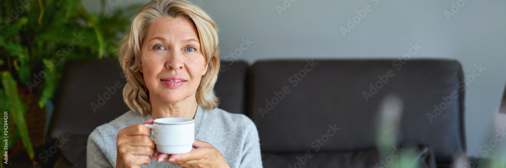 Delicious drink. Cheerful elderly woman sitting on the sofa in her living room and holding a cup of coffee Delicious drink. Cheerful elderly woman sitting on the sofa in her living room and holding a cup of coffee