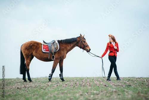 red-haired jockey girl in a red cardigan and black high boots with a horse © omelnickiy