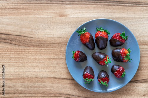 Top view of fresh healthy strawberries dipped in dark chocolate on a blue plate with a wooden background