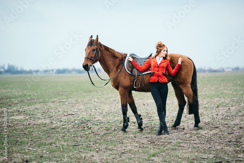 red-haired jockey girl in a red cardigan and black high boots with a horse