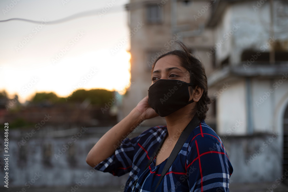 Close up back light portrait of an Indian young woman with corona preventive mask on a rooftop in back-light in home isolation.Indian lifestyle, disease and home quarantine. Close up back light portrait of an Indian young woman with corona preventive mask on a rooftop in back-light in home isolation.Indian lifestyle, disease and home quarantine.