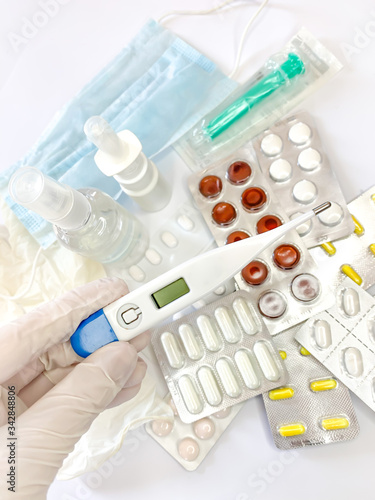 Digital thermometer in hand in glove of a woman on white background with many pills and medicines