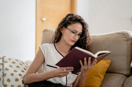 Mujer joven con gafas leyendo un libro con interés desde casa