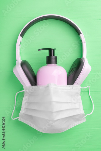 Plastic pink gel soap container with wireless headphones covered with a medical protective mask on a light green wooden background