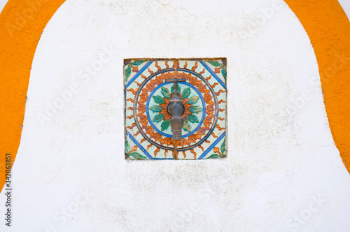 Old copper water faucet, on the white wall with decorative multicolored tile. Spain