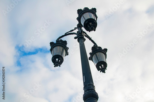 Grey lamppost with three lanterns, cloudy sky. View from the bottom to the top