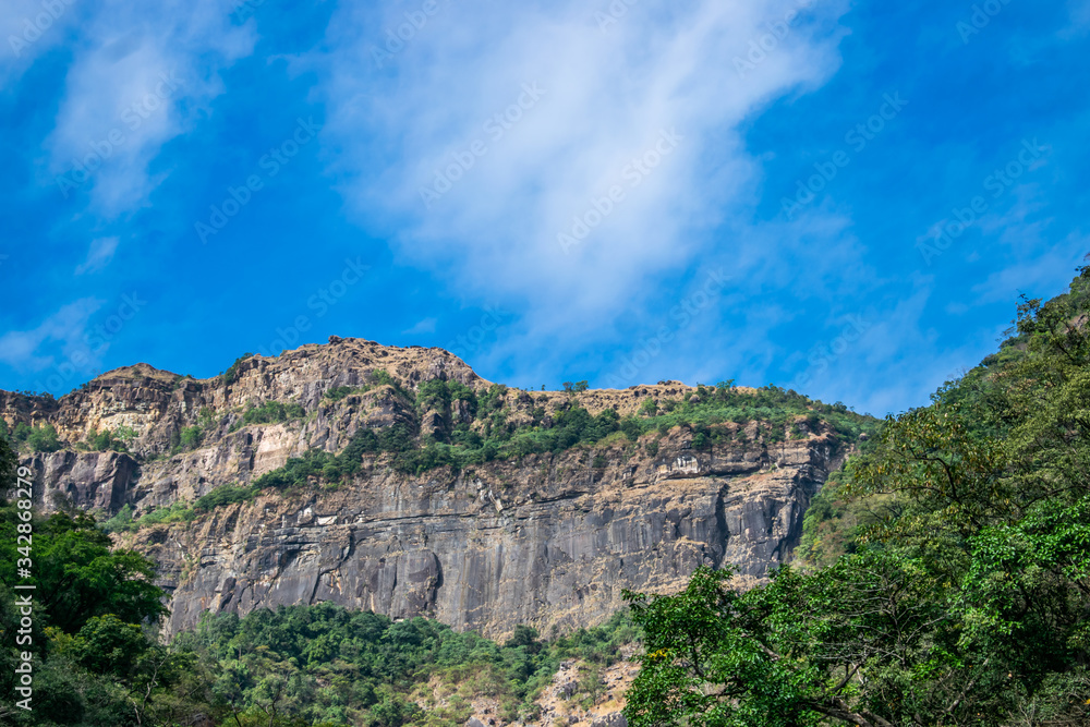Fototapeta premium A huge rocky straight mountain standing tall in the indian state of himachal pradesh. Clear blue skies and brown rocky hill creates a majestic landscape. Snow white cloud in beautiful blue sky.