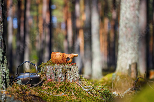 A wooden mug kuksa with a teapot stand on a stump in the forest. Blurred background.