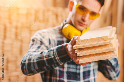 Obraz na płótnie Young professional male joiner in protective goggles, headphones holding wooden planks in hands while working at sawmill factory