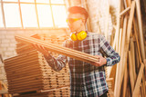 Young skilled carpenter in protective eyewear and headphones working with plywood material at sawmill factory warehouse. Professional male cabinet maker at woodworking workshop. Man artisan, DIY