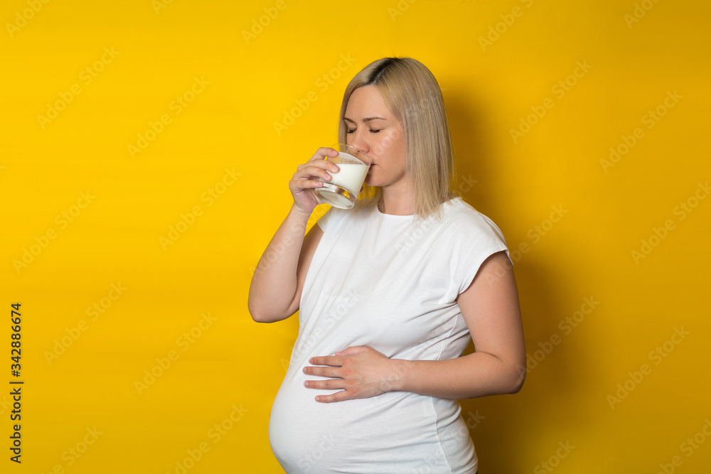 pregnant woman, blonde drinking milk from a glass, holding her hand on her stomach, on a yellow background pregnant woman, blonde drinking milk from a glass, holding her hand on her stomach, on a yellow background