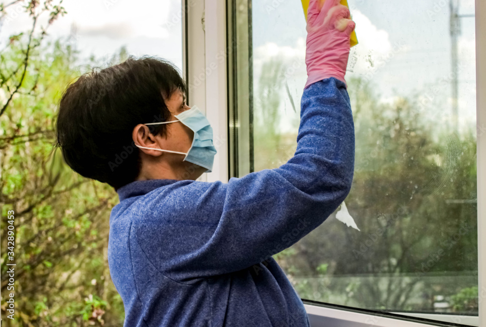 Woman on self-isolation in a blue jacket, latex gloves and a disposable medical mask washes the window with soapy foam after winter. Woman on self-isolation in a blue jacket, latex gloves and a disposable medical mask washes the window with soapy foam after winter.