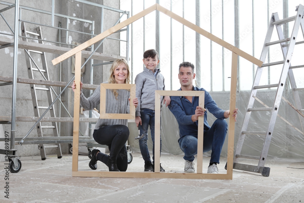 New building residential house purchase apartment concept. Happy family with child hold with their hands the wooden house figure frame with door, window and roof. Inside of interior construction site