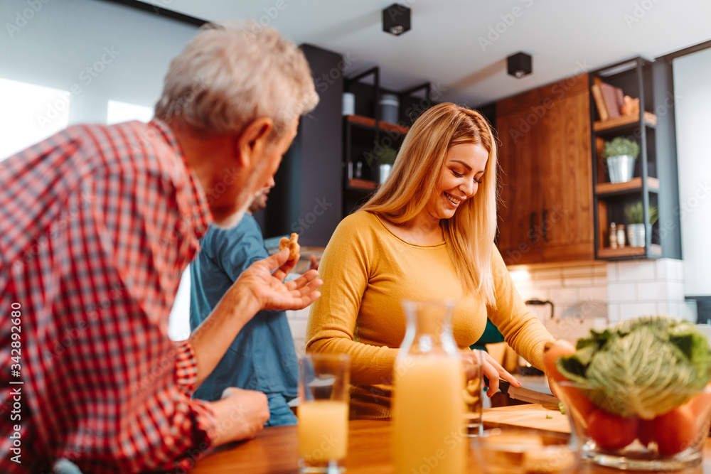Blonde woman is chopping vegetables for salad while her father standing next to her eating pretzels and drink freshly squeezed juice. Blonde woman is chopping vegetables for salad while her father standing next to her eating pretzels and drink freshly squeezed juice.