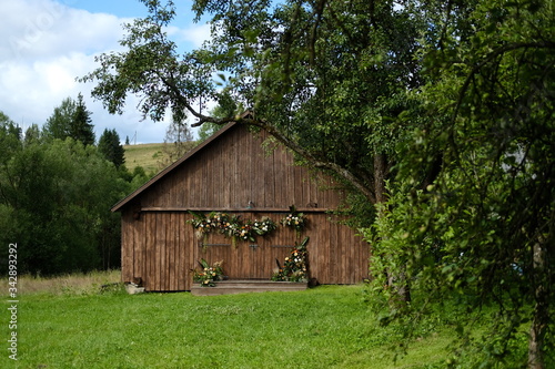 Old barn brown wall wood background surrounded by green trees. The barn is decorated with flowers and greenery. Bright picture of the countryside.