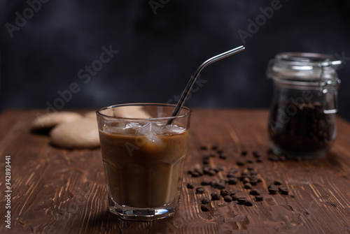 Glass of cold brew iced coffee with milk on brown wood table with coffee beans and cookies on black background