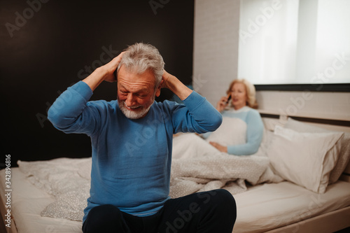 Portrait of elderly man sitting on the bed in bedroom and holding his head because he has headache.