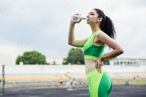 Curly-haired brunette playing sports at the stadium. Girl drinks water from a bottle