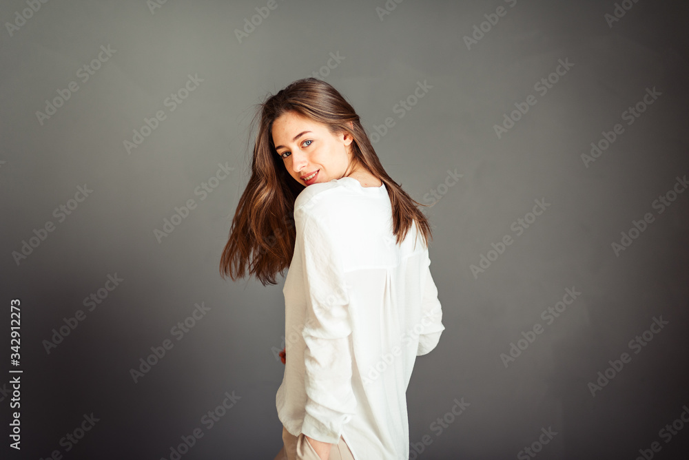 Young girl in a white shirt, sideways hands in a pocket on a gray background. No retouching. Without makeup. Young girl in a white shirt, sideways hands in a pocket on a gray background. No retouching. Without makeup.