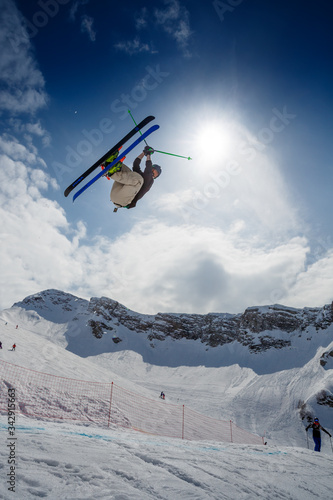 Freestyle ski rider making a spectacular stunt in mountain snow park, ski crossed, against blue sky, mountains, sun and clouds. Extreme winter sport
