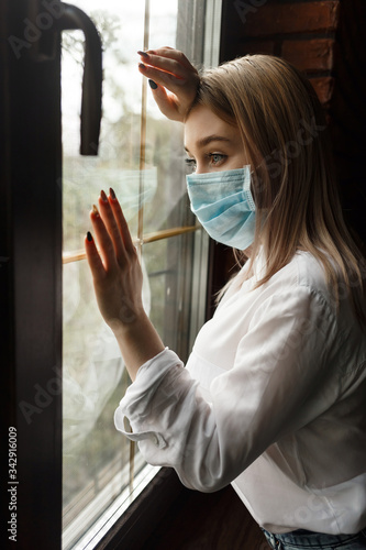 young blonde girl in a protective mask in a dark room looks out the window. Self-isolation from the coronovirus pandemic