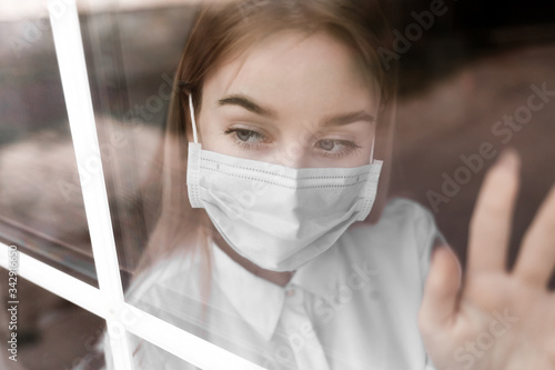 a young blonde girl in a white blouse and a medical mask sits outside the window and looks outside.