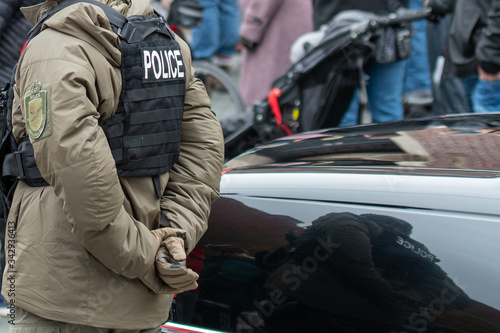 A male police officer wearing a black worn bulletproof vest and a tan colored down filled jacket. The cop is in plain clothes with his hands behind his back. There are people in the background. 