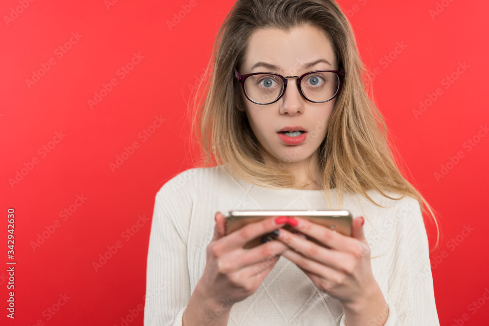 Young girl with glasses with blond hair looks into the smartphone with misunderstanding, stands on a red background Young girl with glasses with blond hair looks into the smartphone with misunderstanding, stands on a red background