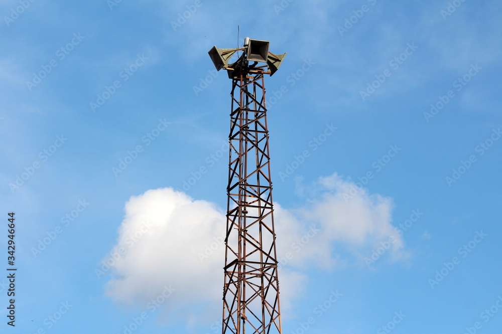 Tall partially rusted metal structure holding four large public civil defence warning air sirens on top on cloudy blue sky background Tall partially rusted metal structure holding four large public civil defence warning air sirens on top on cloudy blue sky background