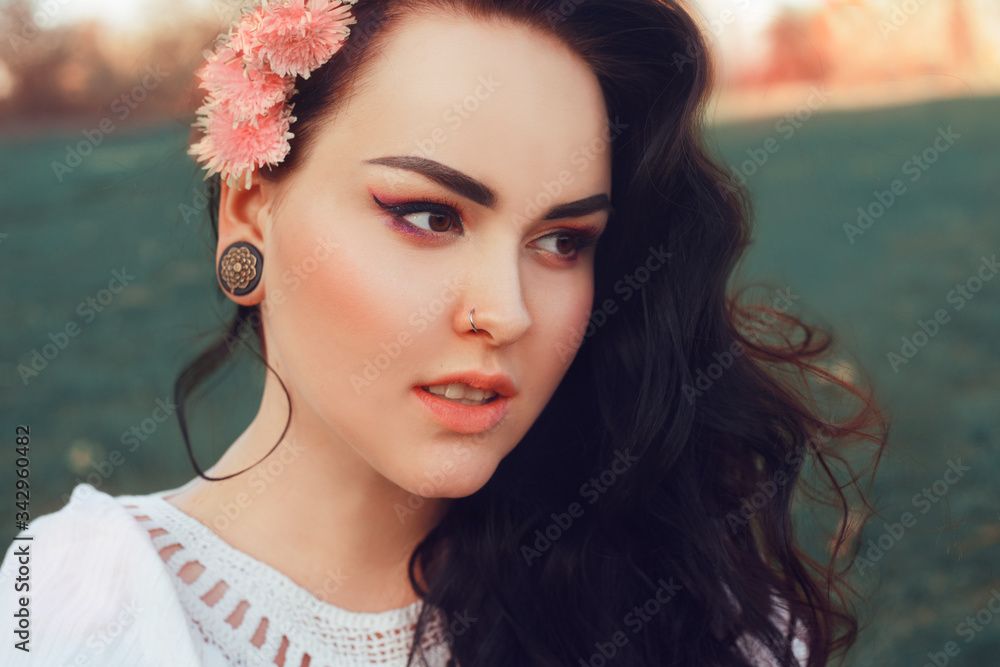 Fototapeta premium Gorgeous brunette sitting in the park on the grass in a white boho dress with pink flowers in her hair in a fashionable finish.