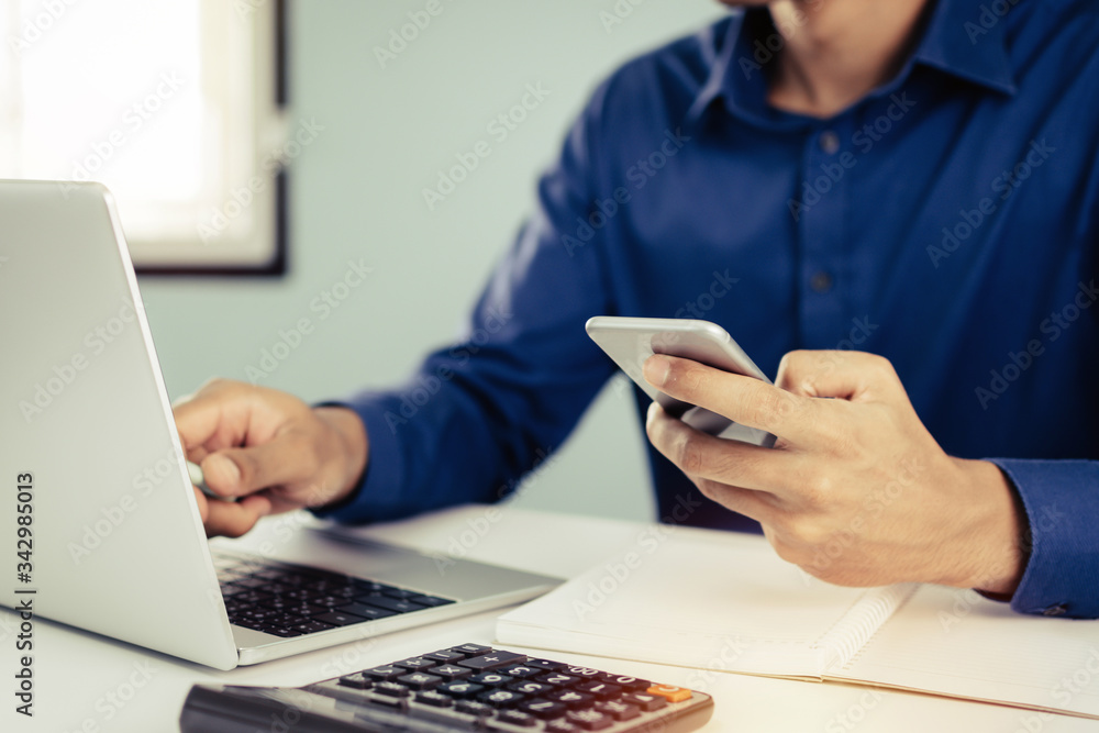 business man in blue shirt using mobile phone working with laptop computer and document report on desk at home, self quarantine, work from home, marketing, business finance, digital technology concept business man in blue shirt using mobile phone working with laptop computer and document report on desk at home, self quarantine, work from home, marketing, business finance, digital technology concept