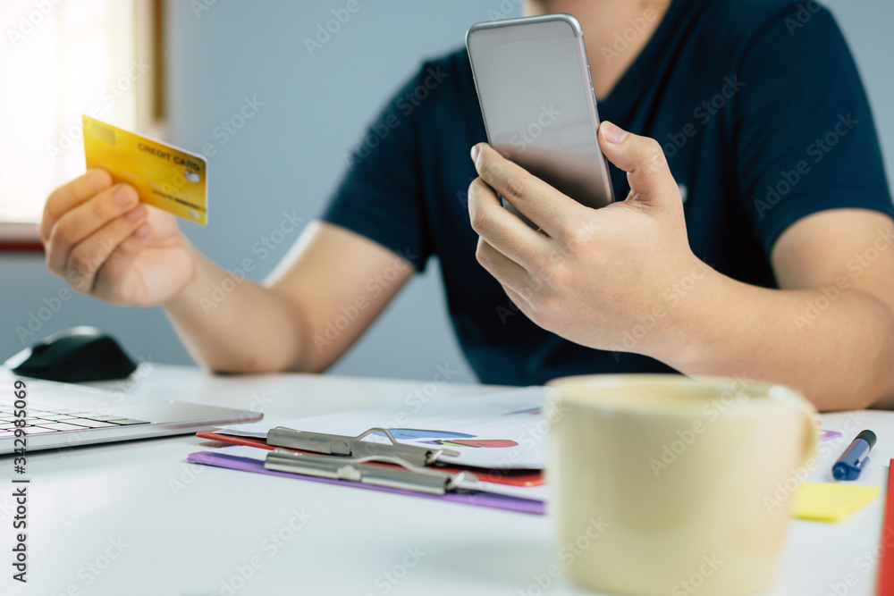 Fototapeta premium young man in blue casual clothes entering security code with mobile phone and paying with credit card and document report on desk at home, shopping online, electronic pay, digital technology concept