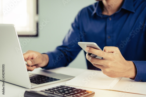 business man in blue shirt using mobile phone working with laptop computer and document report on desk at home, self quarantine, work from home, marketing, business finance, digital technology concept