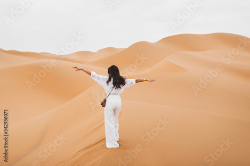 Woman in stylish white shirt and pants with retro photo camera happy to explore Sahara desert sand dunes in Morocco. View from the back.