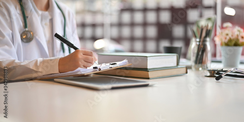 Cropped image of young doctor writing on clipboard while sitting at the doctor working desk that surrounded by stack of books, pencils in glass vase and flowers over orderly workplace as background.
