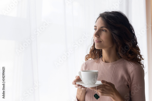 Dreamy young beautiful woman standing near window with cup of tea, enjoying relaxed lazy morning alone at home. Peaceful millennial attractive lady traveler visualizing future, planning day at hotel.