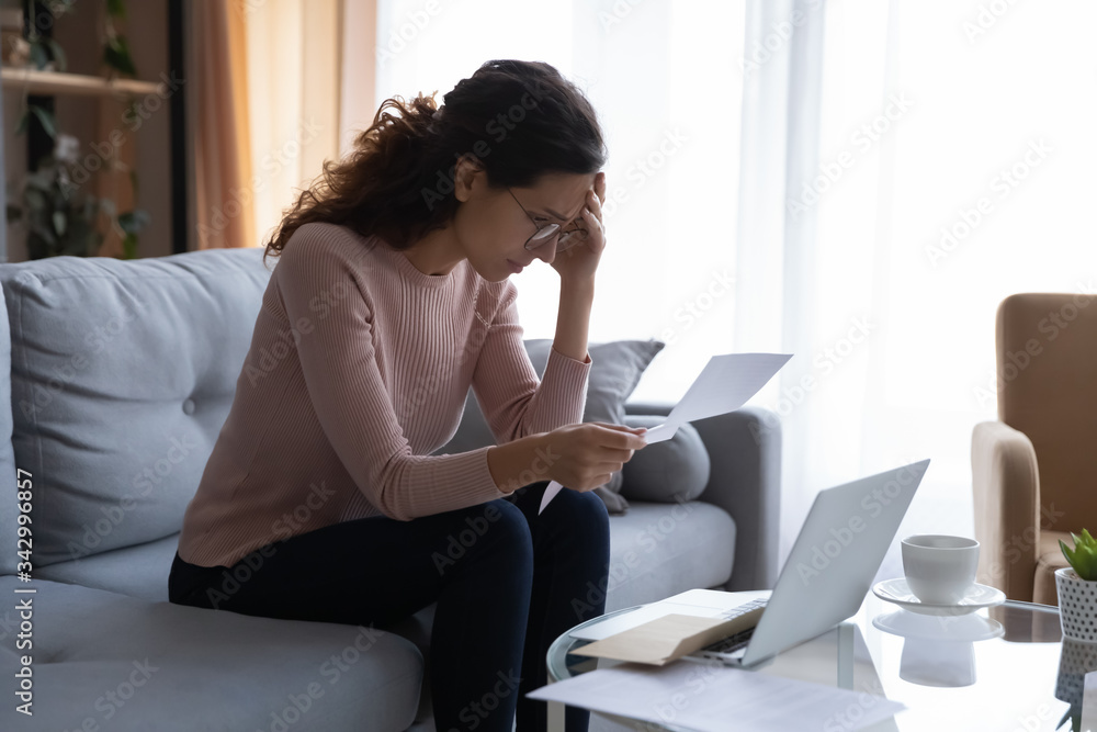 Frustrated desperate young woman in eyeglasses touching forehead, looking through banking credit termination, having financial problems, reading refusal document, feeling depressed alone at home. Frustrated desperate young woman in eyeglasses touching forehead, looking through banking credit termination, having financial problems, reading refusal document, feeling depressed alone at home.
