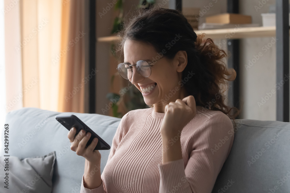 Head shot emotional young woman in glasses looking at mobile screen, amazed by good news notification. Overjoyed pretty lady celebrating online lottery win, reading success victory message at home.