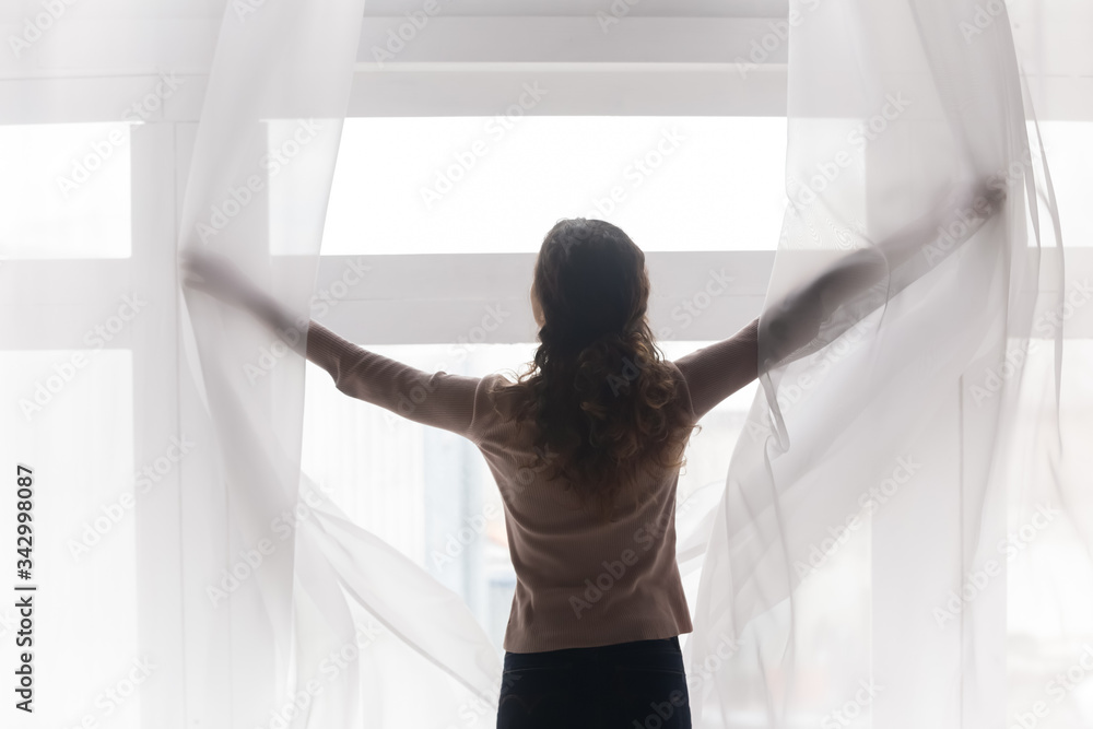 Back rear view happy young woman opening light curtains, welcoming new day. Millennial brunette lady pulling lace in morning, enjoying morning panoramic view from huge window at home or hotel. Back rear view happy young woman opening light curtains, welcoming new day. Millennial brunette lady pulling lace in morning, enjoying morning panoramic view from huge window at home or hotel.